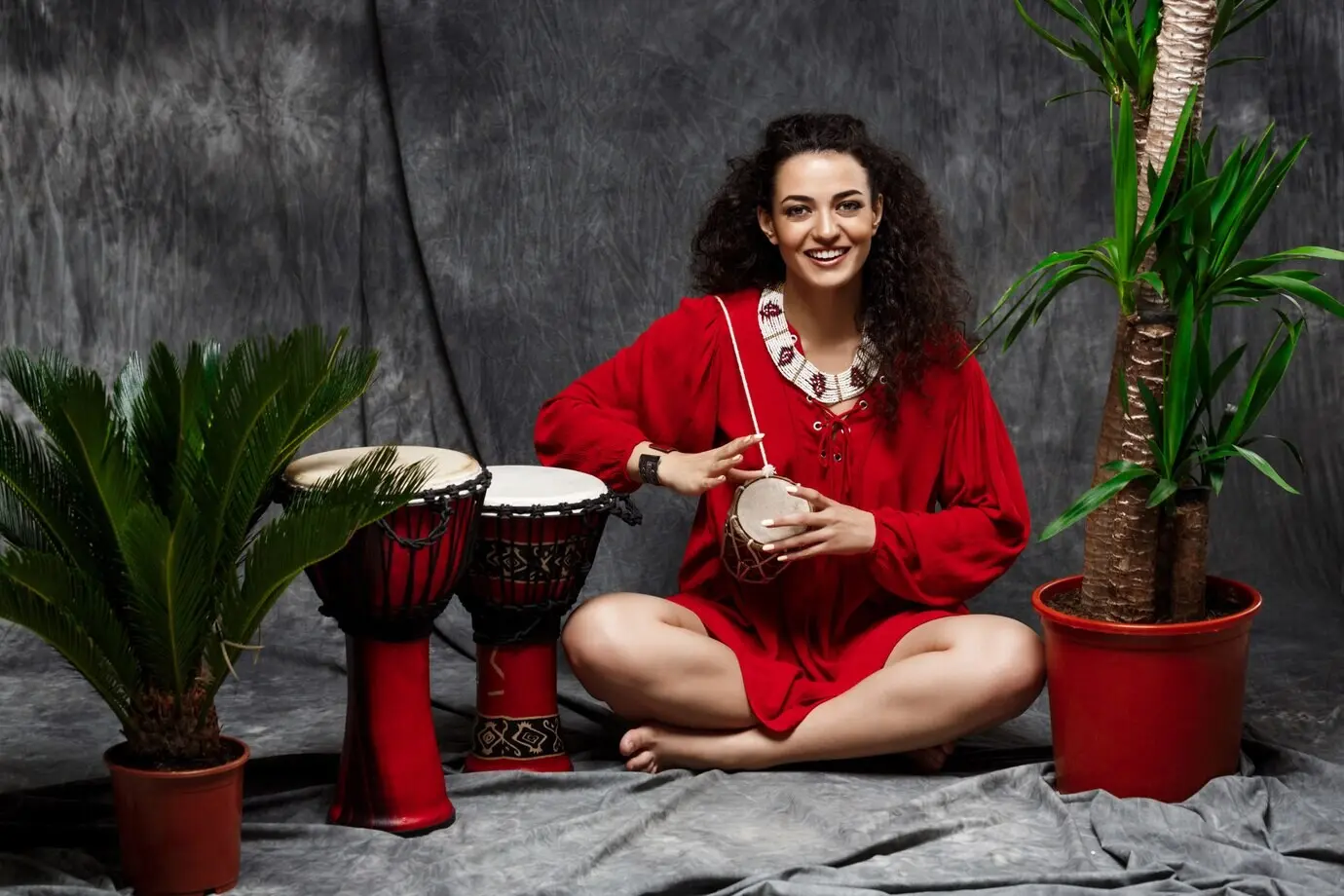 Schönes Mädchen spielt eine Trommel inmitten tropischer Pflanzen vor einer grauen Wand.